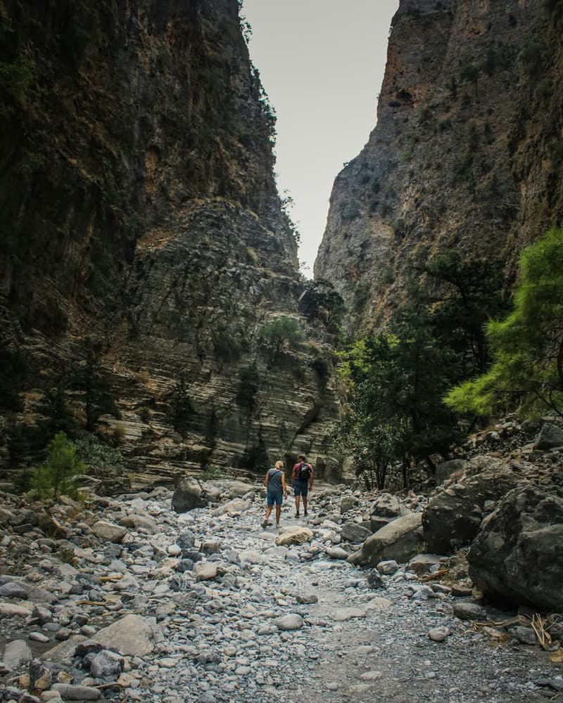 Two hikers walking a dry riverbed through a narrow limestone gorge.