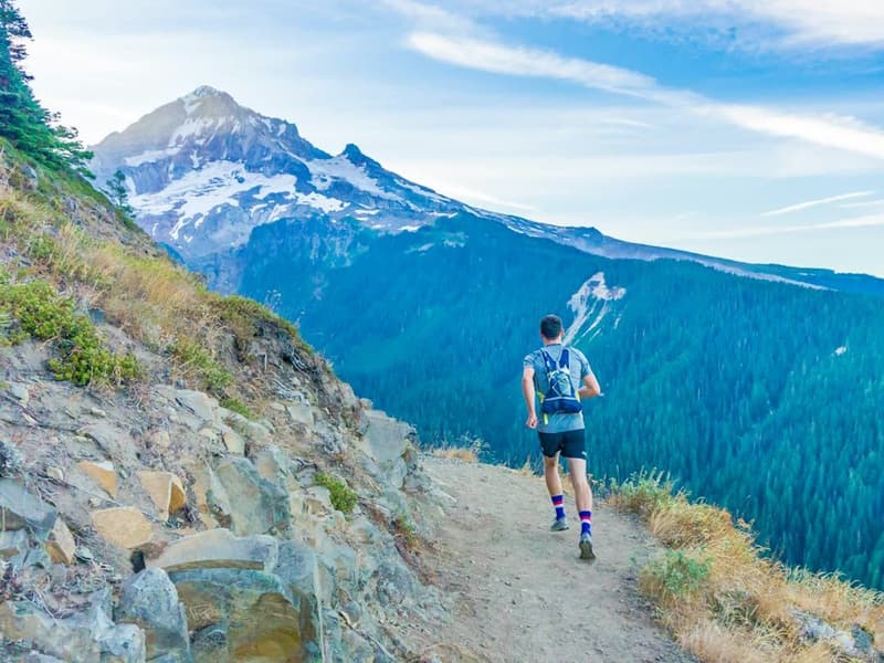 A trail runner on a mountain ridge path below a snowy peak.